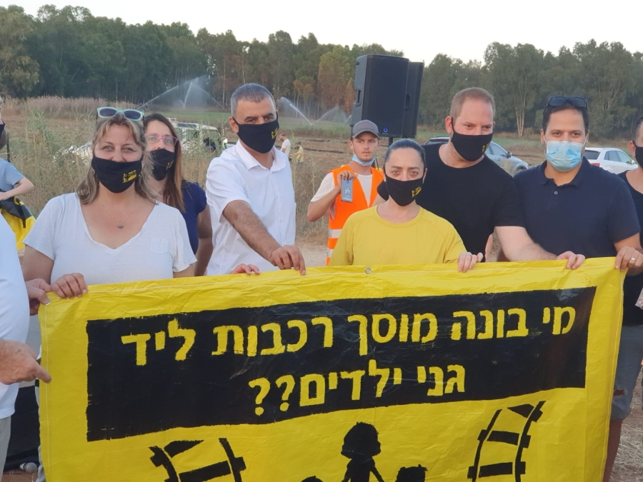 משתתפים בהפגנה מחזיקים שלט עם הכיתוב מי בונה מוסך רכבות ליד גני ילדים